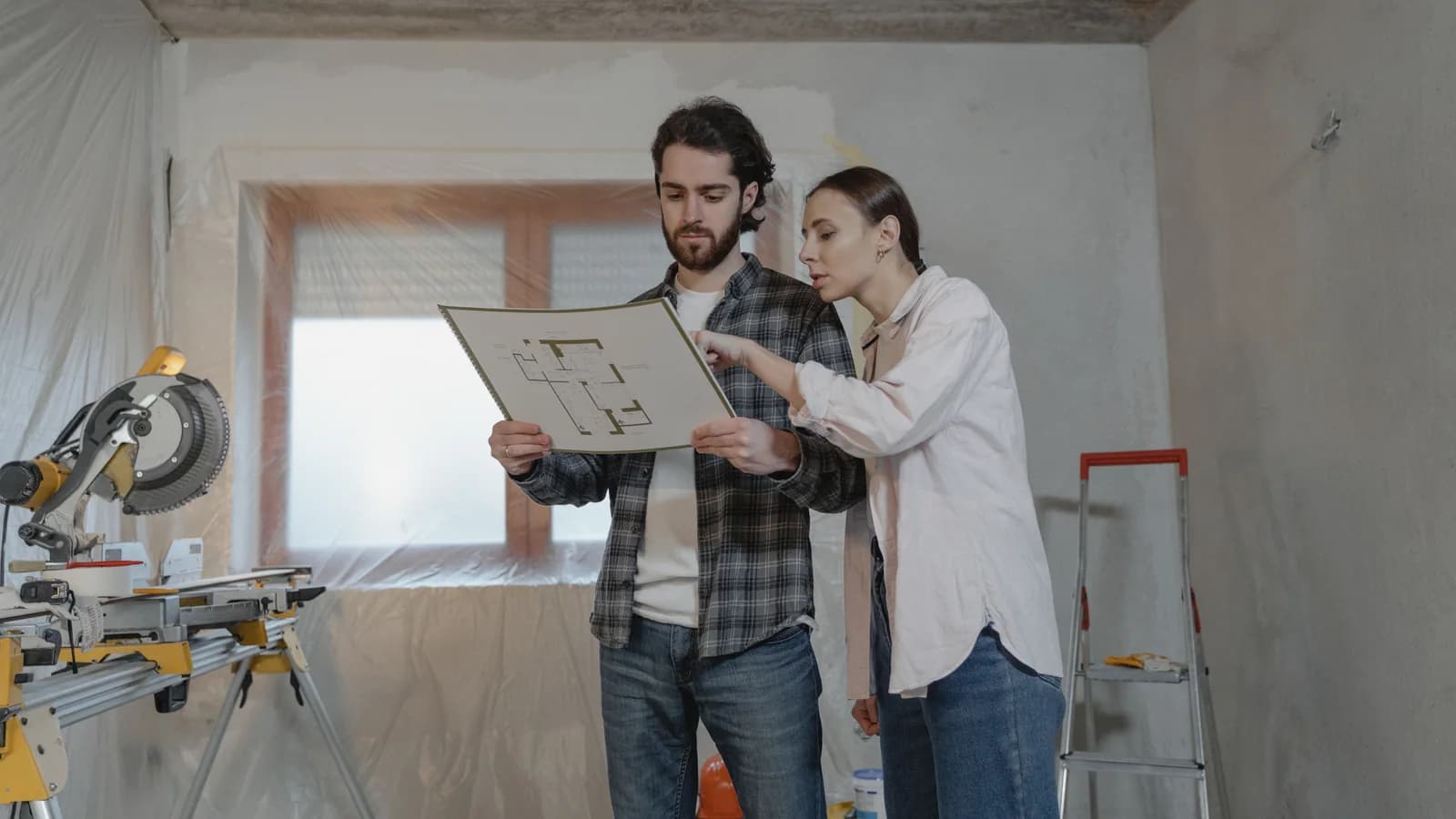Two people reviewing a floor plan in a room under renovation